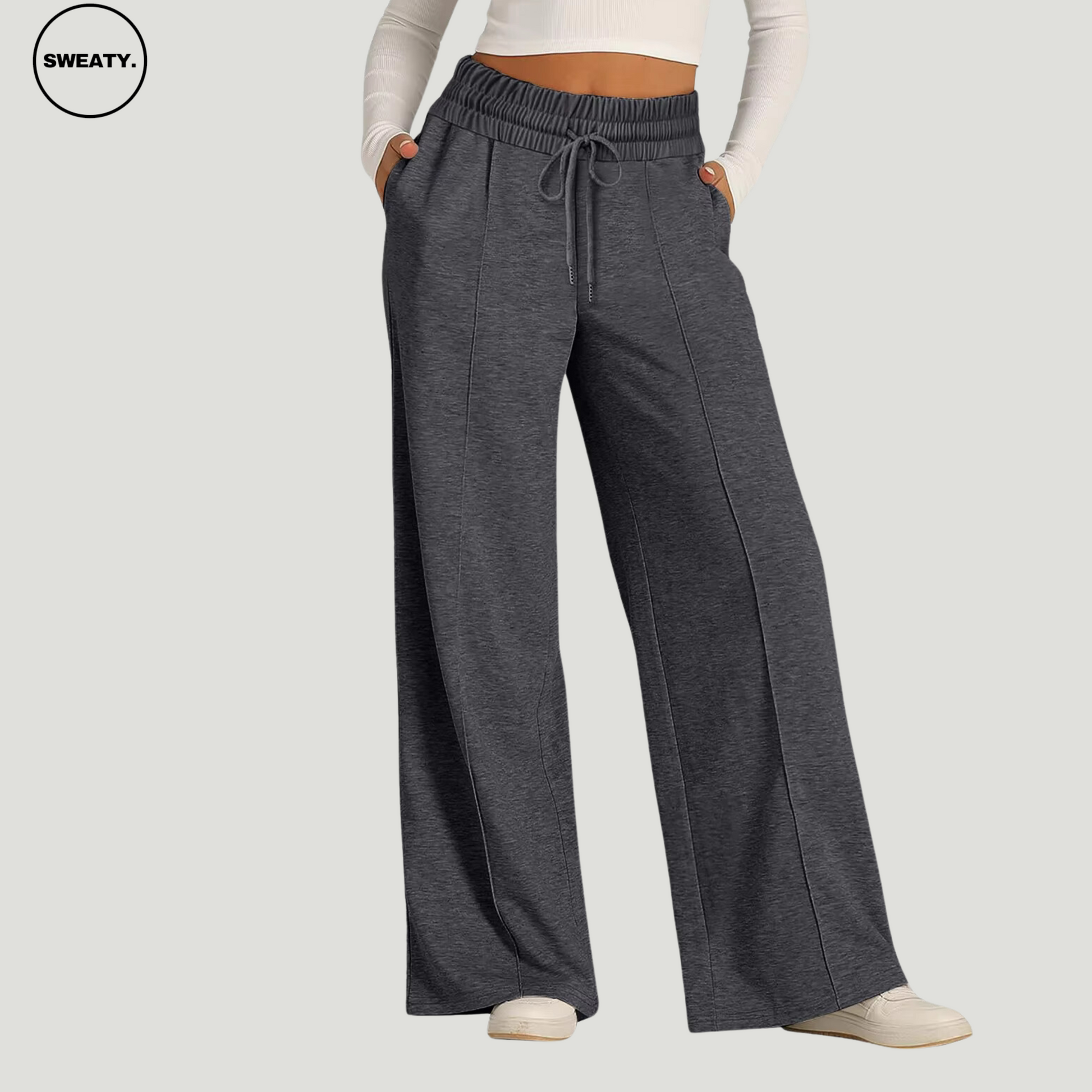 Front view of a woman wearing dark grey wide-leg fleece-lined sweatpants, featuring an elastic drawstring waistband and side pockets. These relaxed-fit pants provide comfortable wear for lounging or casual outings. Paired with a white long-sleeve crop top and white sneakers.