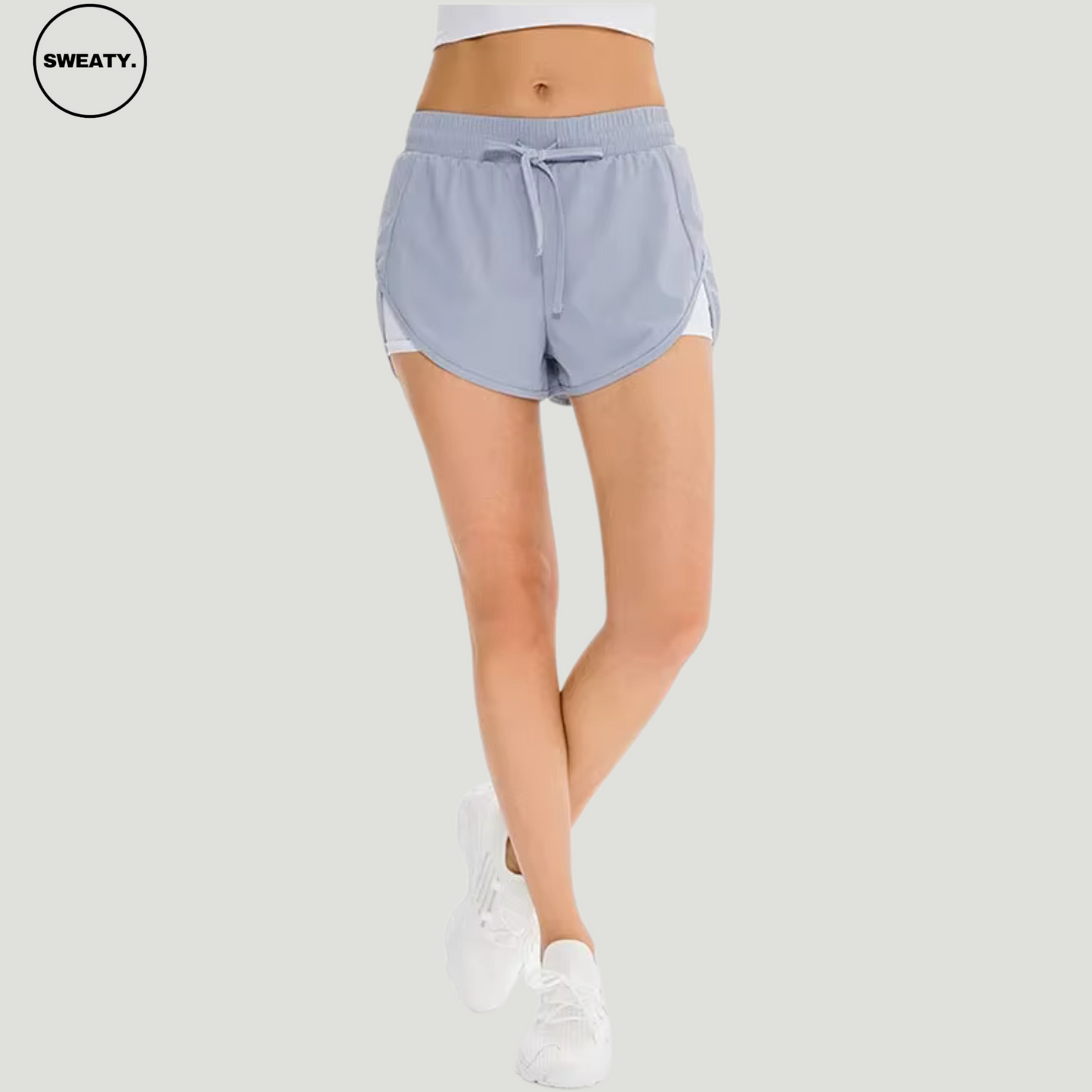 Front view of a woman wearing a light blue 2-in-1 skort by SWEATY, featuring a drawstring waistband and built-in white shorts for added support. The skort is designed for activewear, with a sporty and stylish look. Paired with white athletic shoes, this outfit offers a sleek and practical design for workouts or casual activities.