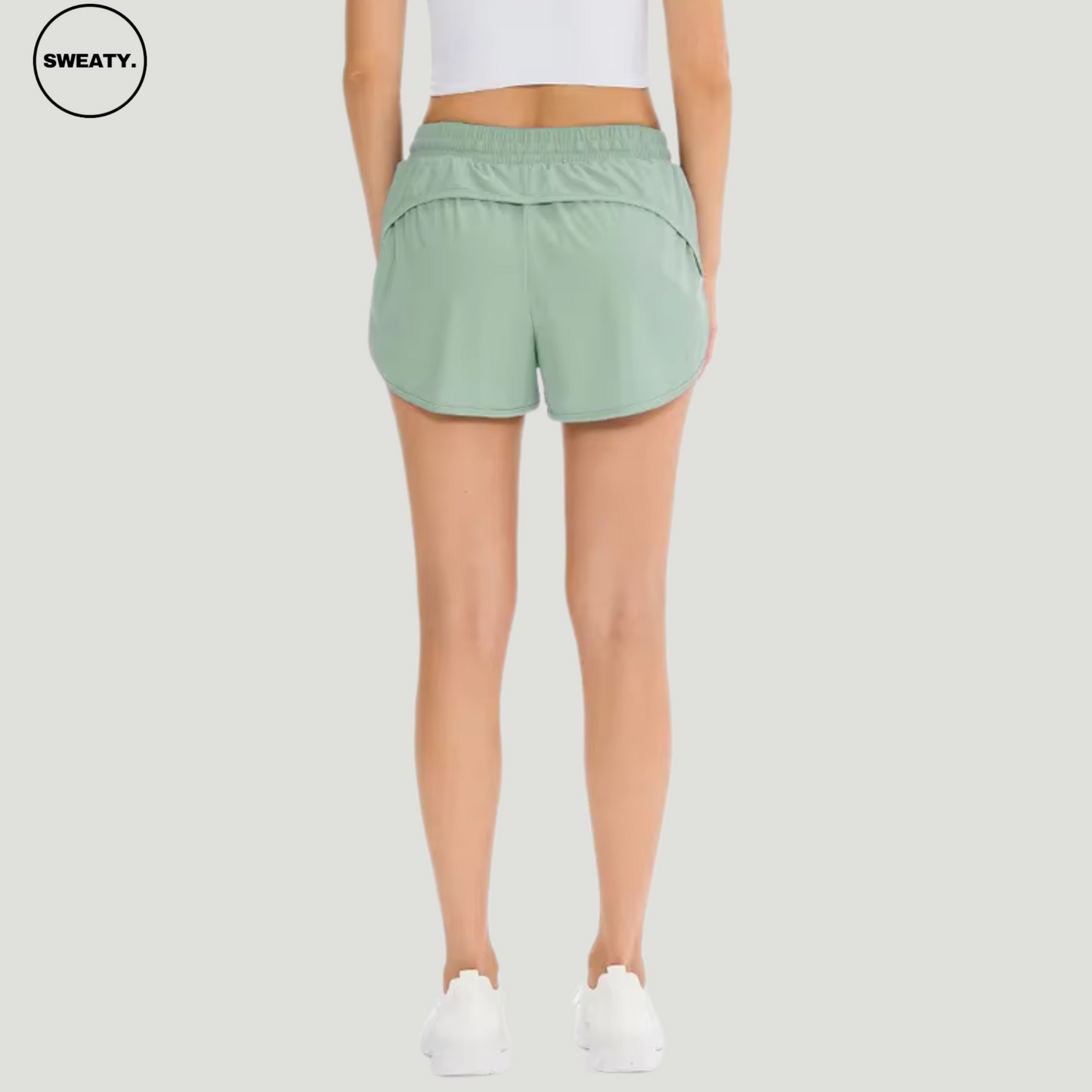 Back view of a woman wearing a mint green 2-in-1 skort by SWEATY, featuring an elastic waistband and layered design for added coverage. The skort provides a comfortable and sporty fit, ideal for active wear. Paired with white sneakers and a white crop top.